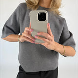 En person iført en grå bluse tager et billede med en smartphone. Personen står i et indendørs, neutralt oplyst rum og bærer et armbånd.