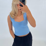 Person tager et spejlbillede med en smartphone. De har en blå top, sort bukser og gyldent smykke på. Baggrunden er helt hvid, hvilket skaber en minimalistisk stil.