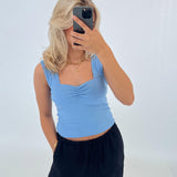 Person tager et selfie iført en lyseblå top og sorte bukser. De står mod en hvid baggrund og bærer en guldhalskæde og armbånd, mens de dækker ansigtet med en telefon.