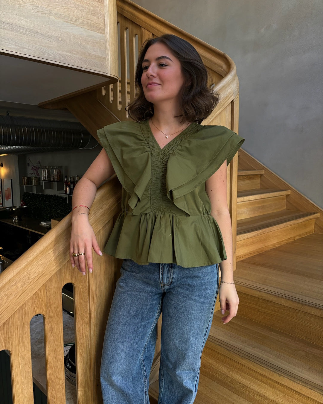 Kvinde står på trappe, iført en grøn bluse og blå jeans. Hun hviler hånden på gelænderet i en moderne, træinteriørindretning med dæmpet belysning.