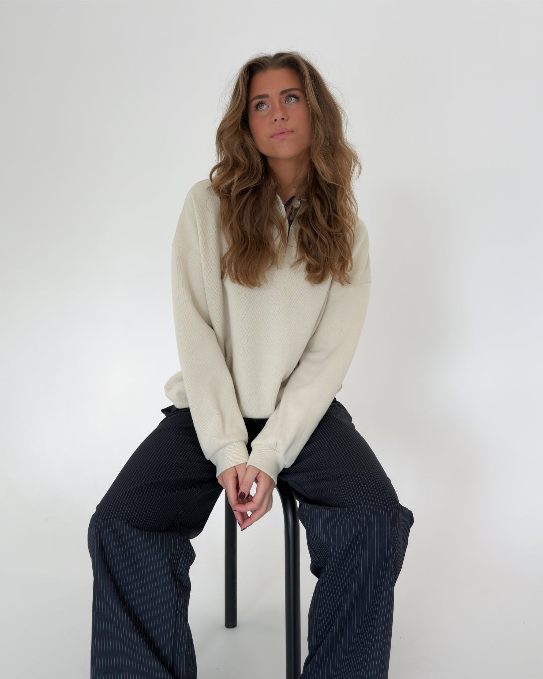 En person sidder på en stol med samlet hænder, iført en beige trøje og mørke bukser. Baggrunden er lys og minimalistisk.