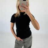 En person klædt i sort t-shirt og grå jeans tager et spejlbillede med en smartphone i en lys baggrund. En hånd hviler i bukselommen.