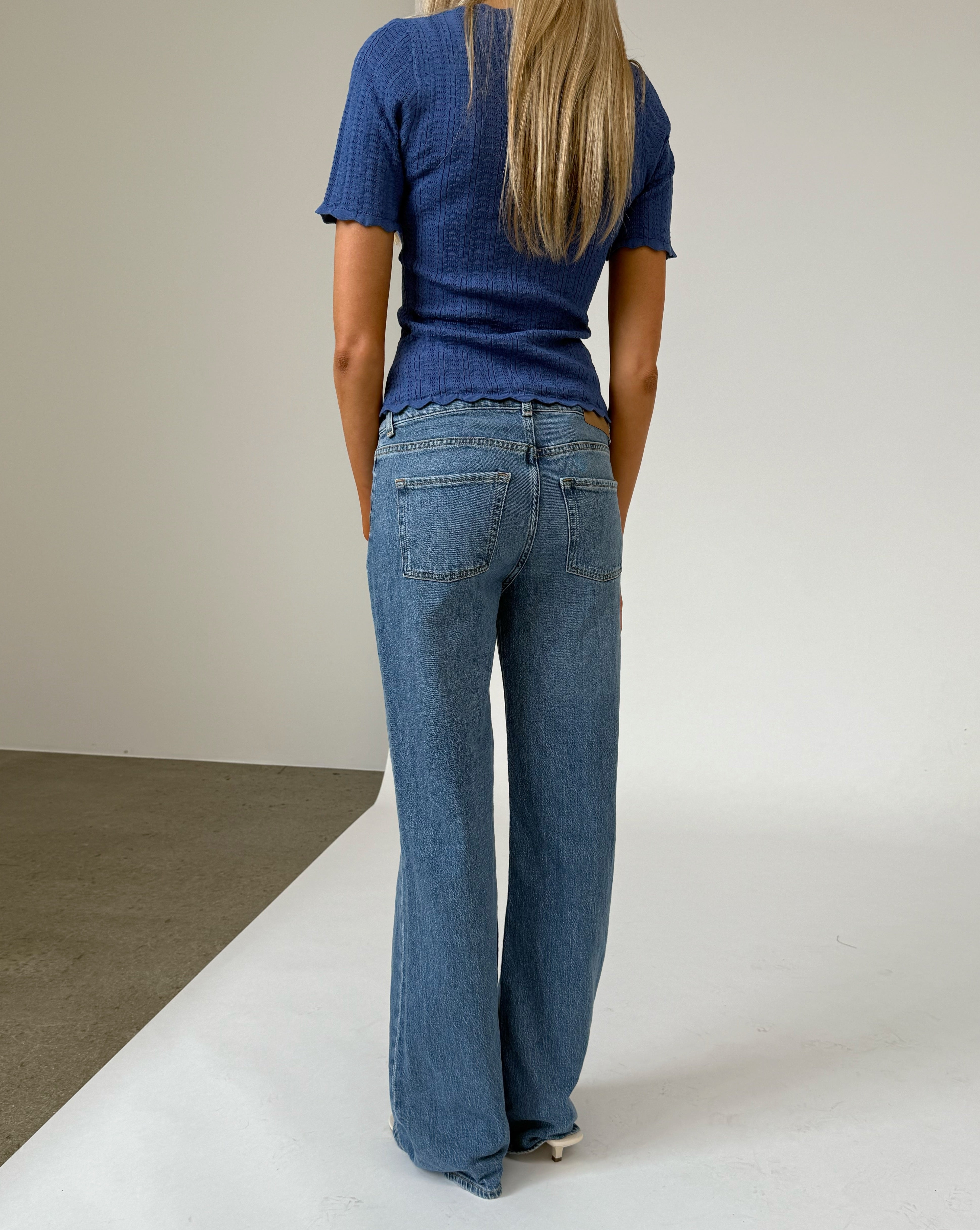En kvinde i en blå bluse og jeans står med ryggen til i et neutralt, indendørs miljø med en lys væg og betonlignende gulv.