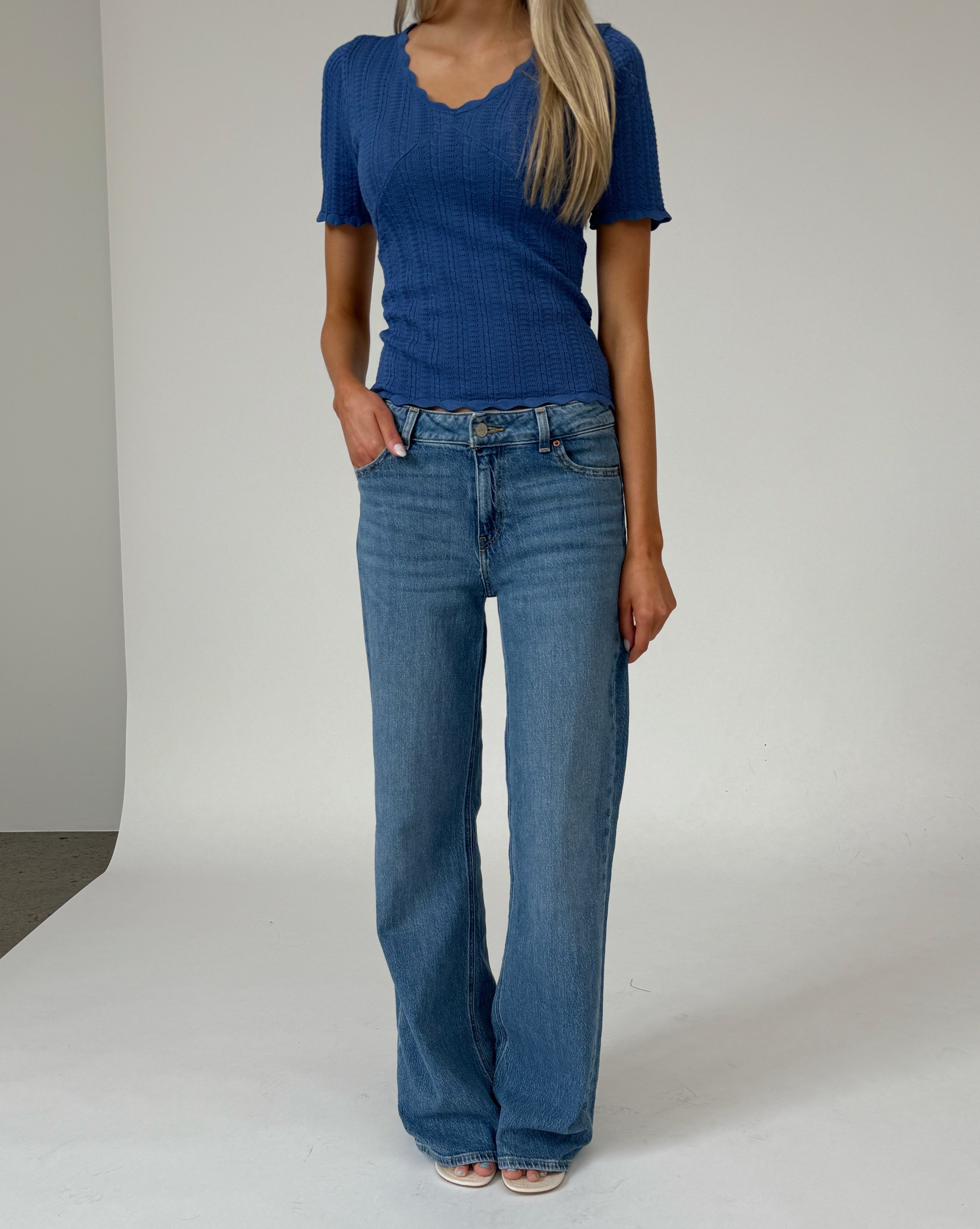 En person står iført en blå kortærmet bluse og blå jeans i et minimalistisk studie med en neutral baggrund.