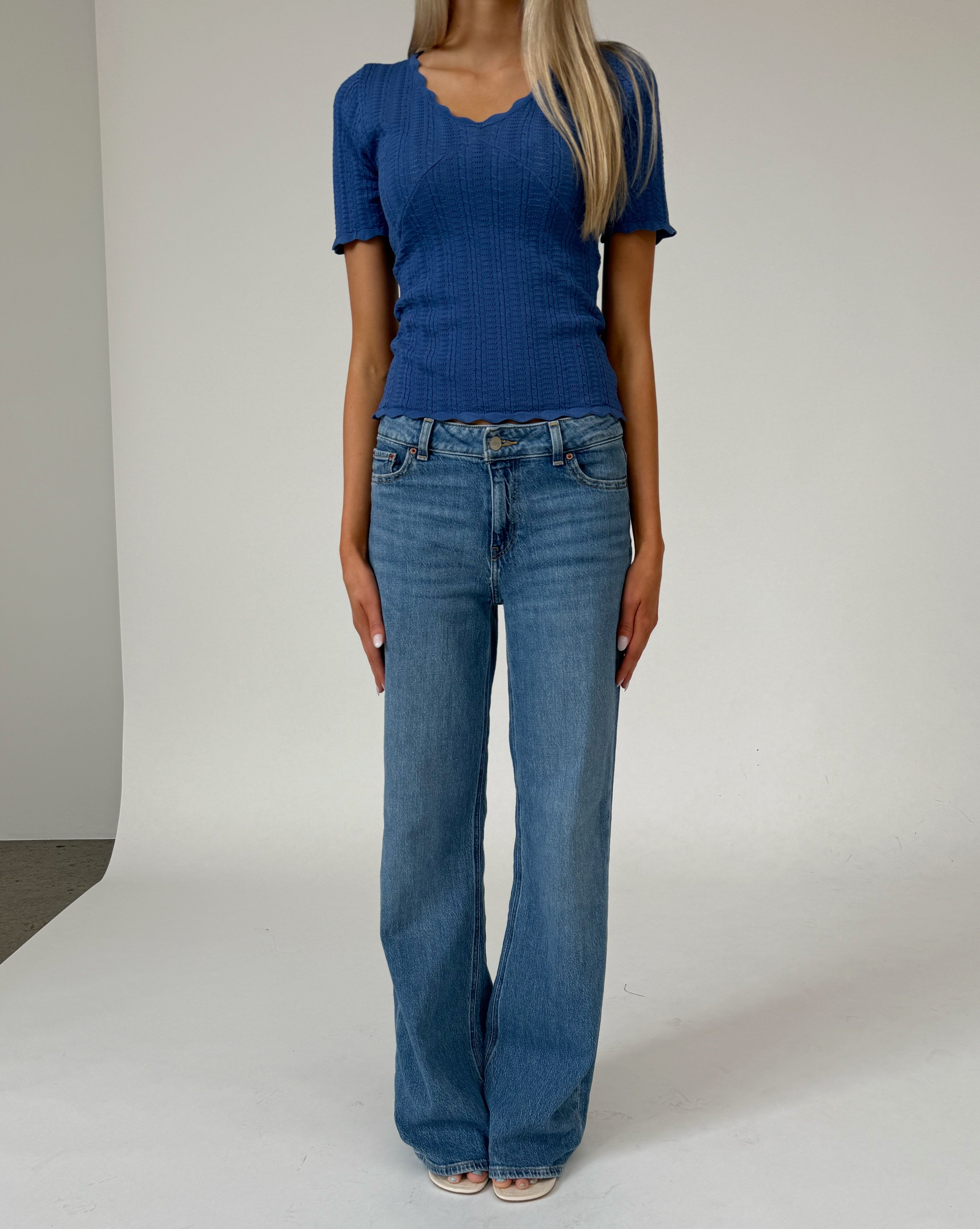 En person står i midten iført en blå kortærmet trøje og blå jeans. Baggrunden er en lys, neutral indendørs væg uden tekst.