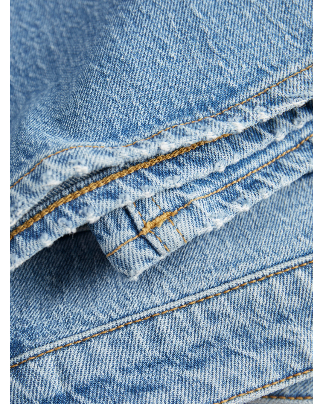 Blå denimstof ligger foldet med synlige gulbrune syninger. Stoffet er ru med let slidte kanter, hvilket er typisk for jeansmateriale.