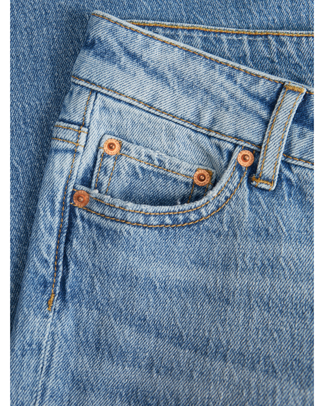 Et par blå jeans ligger fladt, med fokus på lommen dekoreret med kobbernitter. Synlige sømme og detaljer viser stoffets struktur i et enkel og tidløst design.