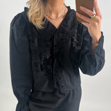 En person i sort bluse med flæsedetaljer tager en selfie med en smartphone. Personen har lyst hår og bærer minimal smykker i et neutralt oplyst rum.