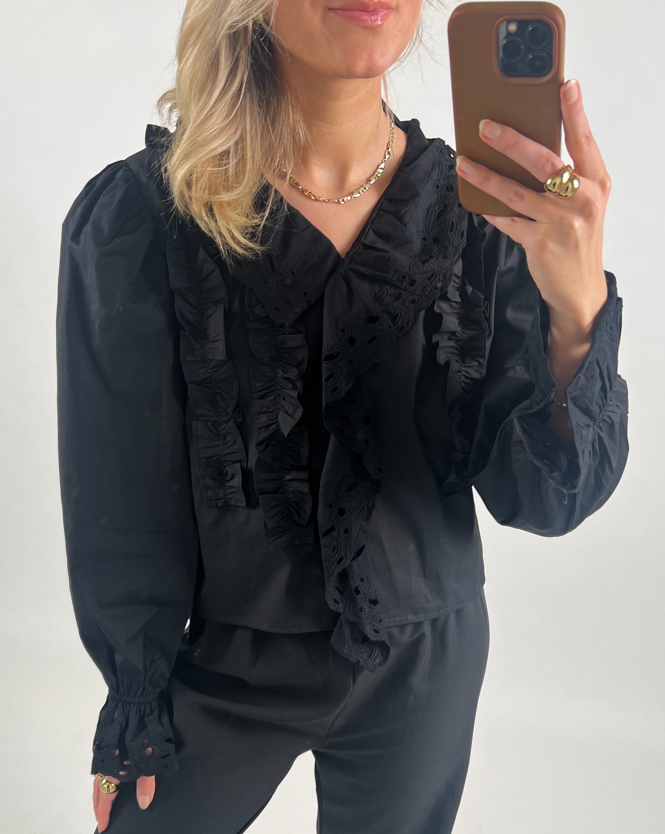 En person i sort bluse med flæsedetaljer tager en selfie med en smartphone. Personen har lyst hår og bærer minimal smykker i et neutralt oplyst rum.