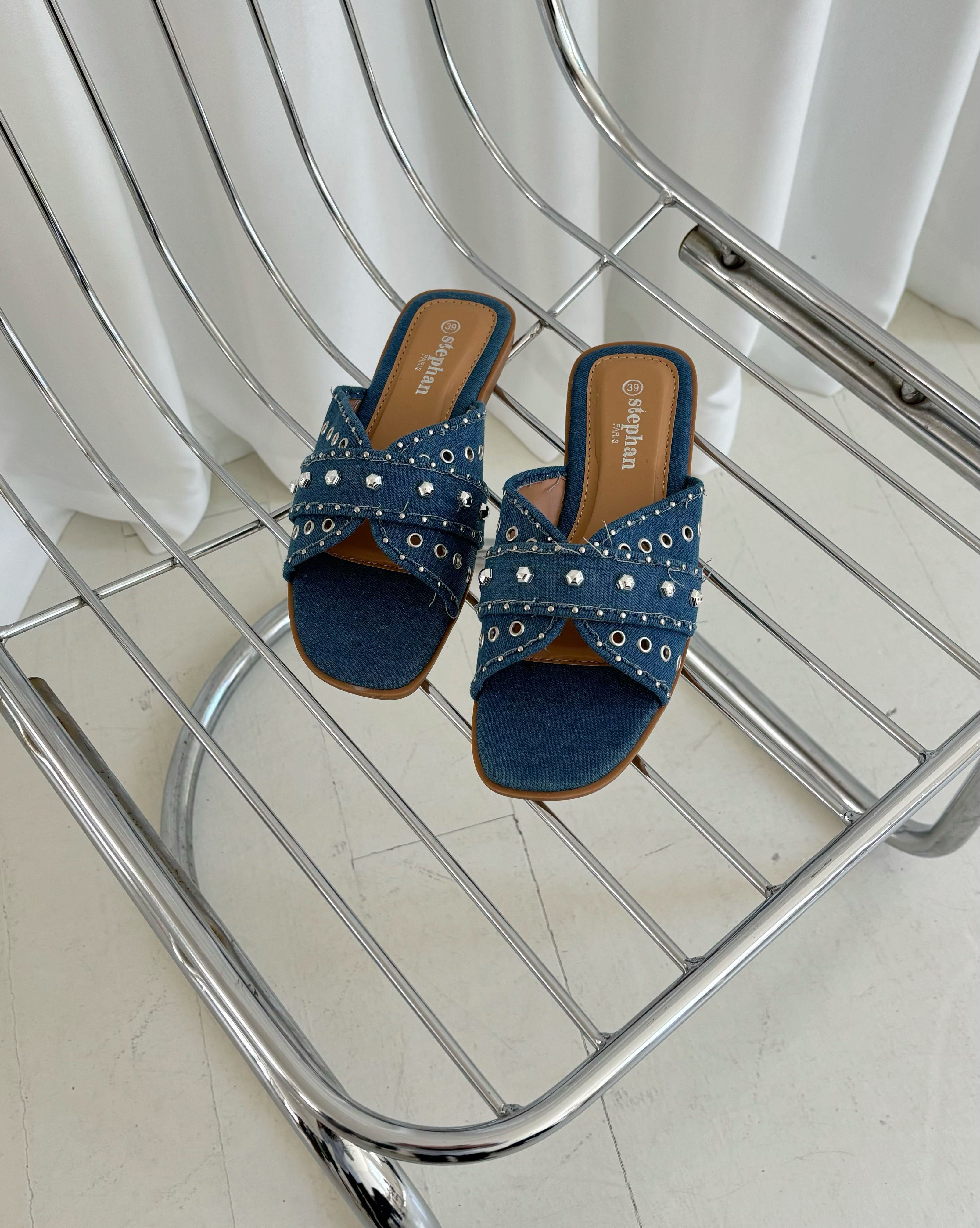 Sorte denim-sandaler med nitter ligger stille på en moderne metalstol. Tekst på indersålen: "stephan shoes." Gardiner i baggrunden skaber en rolig atmosfære.