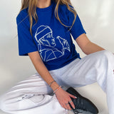 En person sidder afslappet iført en blå t-shirt med en hvid lineart illustration. De bærer hvide bukser og sorte sneakers. Baggrunden er neutral.