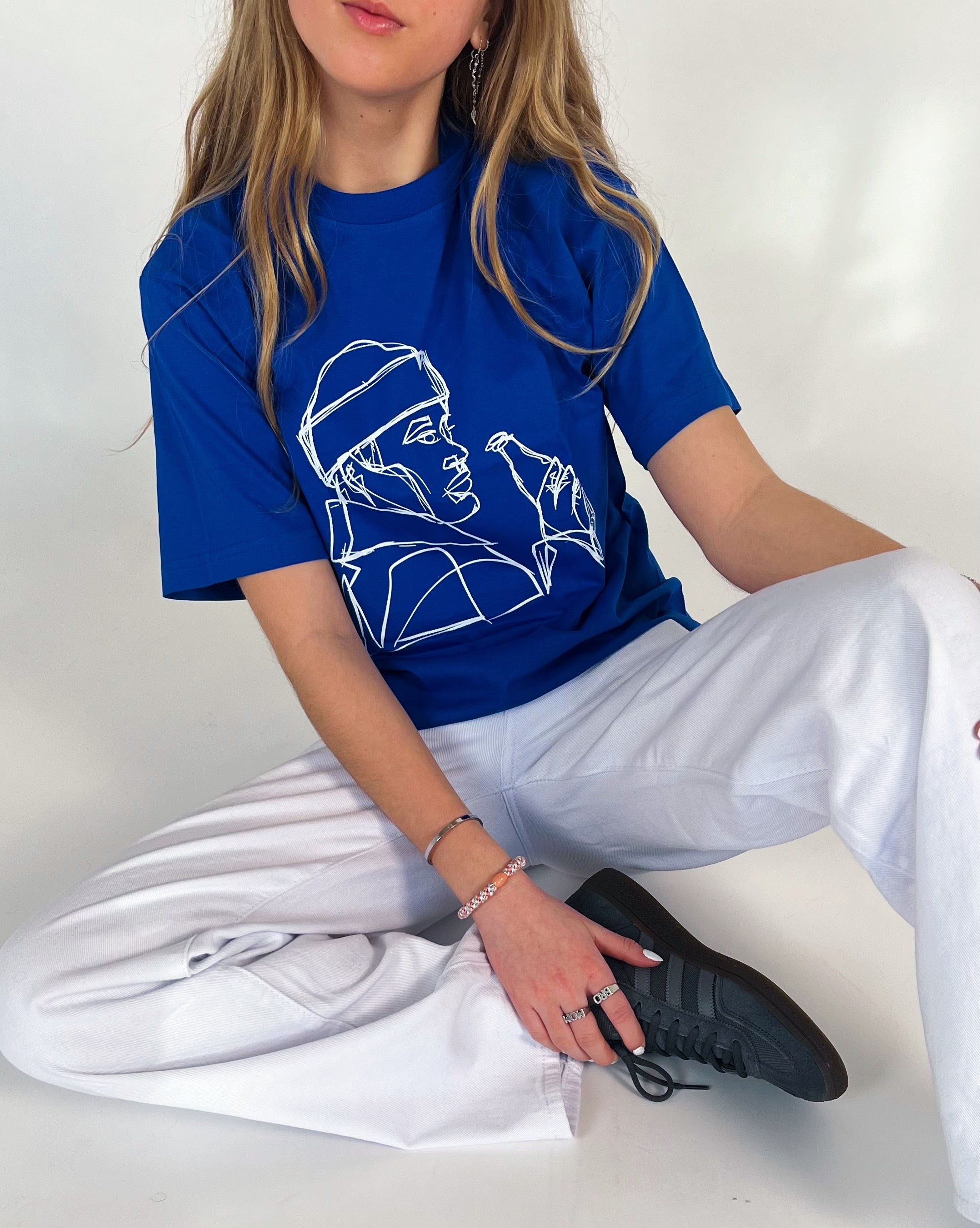 En person sidder afslappet iført en blå t-shirt med en hvid lineart illustration. De bærer hvide bukser og sorte sneakers. Baggrunden er neutral.