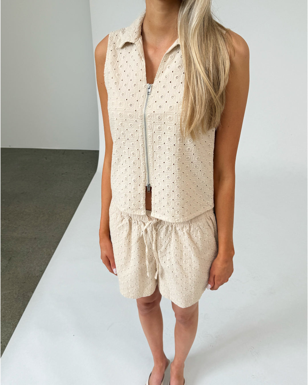 En kvinde står iført en beige, ærmeløs top og shorts med et mønster af små huller. Hun er i et indendørsmiljø med en hvid baggrund og gråt gulv.