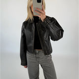 En person iført en sort læderjakke tager et spejlfoto med en smartphone i et minimalistisk, hvidt rum. Personen har lyse hår og grå jeans på.