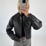 En person tager et spejlbillede, iført en sort læderjakke og grå jeans, i et lyst rum. Personen holder en mobiltelefon foran ansigtet.