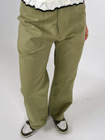 Olivenfarvede bukser bæres af en person, der står op, kombineret med grønne og hvide sneakers og en hvid strikket trøje med sort kant. Baggrunden er hvid.