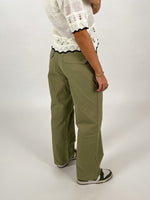 En person iført grønlige bukser og en hvid, strikket bluse står sidelæns på en lys baggrund, med hænderne afslappet ved siden.