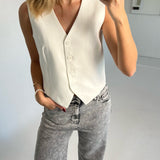 En person med lyseblondt hår bærer en hvid vest og stenvasket jeans, mens de tager en selfie i et minimalistisk rum. Personen har hånden i lommen og bærer et armbånd.