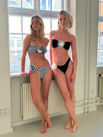 To kvinder i bikinier står oprejst ved siden af hinanden. Den ene bærer en stribet bikini, den anden en sort. De befinder sig i et rum med store vinduer og radiatorer.