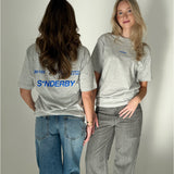 To kvinder iført grå T-shirts står ved siden af hinanden i et neutralt rum. Den ene vender ryggen til, mens den anden ser fremad. T-shirt teksten lyder: "S*NDERBY".