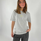 En person iført en grå T-shirt med teksten "S'NOEBY" står afslappet med hånden i lommen på et par mørke bukser, mod en lys baggrund.