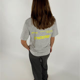 En person med langt hår står med ryggen til iført grå T-shirt og mørke bukser. Tekst på T-shirten: "301103 10-04-2021 BY SOFIE SØNDERBY SNDRBX STUDIO SØNDERBY." Baggrund er enkel og lys.