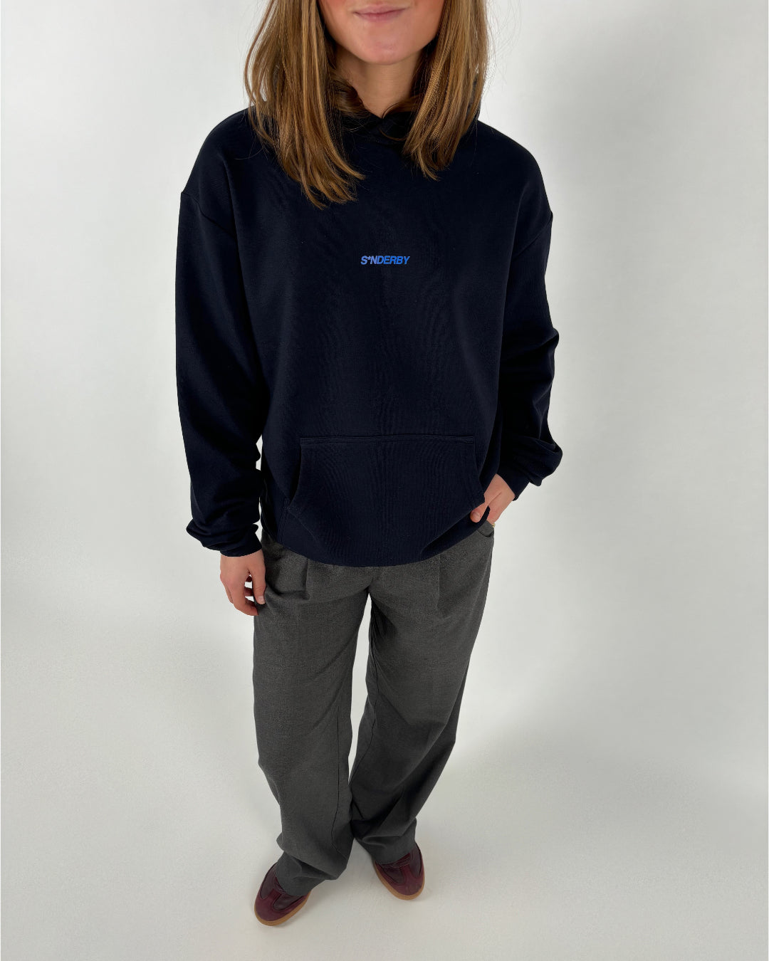En person iført en sort sweatshirt med teksten "SØNDERBY" står i hvilestilling; bukselommerne hviler i lysegrå bukser med brune sko på et hvidt baggrund.