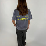 En person står med ryggen til iført en grå T-shirt med gul tekst: "301103 10-04-20-21 BY-SOFFE SØNDERBY SNDB X BS S*NDERBY" og sorte bukser i en lys baggrund.