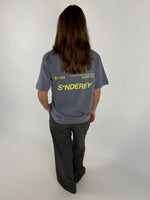 En person står med ryggen til iført en grå T-shirt med gul tekst: "301103 10-04-20-21 BY-SOFFE SØNDERBY SNDB X BS S*NDERBY" og sorte bukser i en lys baggrund.