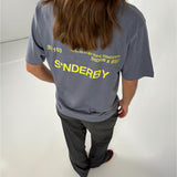 En person med langt hår står i et hvidt rum, iført en grå T-shirt med gul tekst: "30 / 103, 16-04-20-21-BY-SOFIE-SØNDERBY, SNDRB X BSS, S*NDERBY".