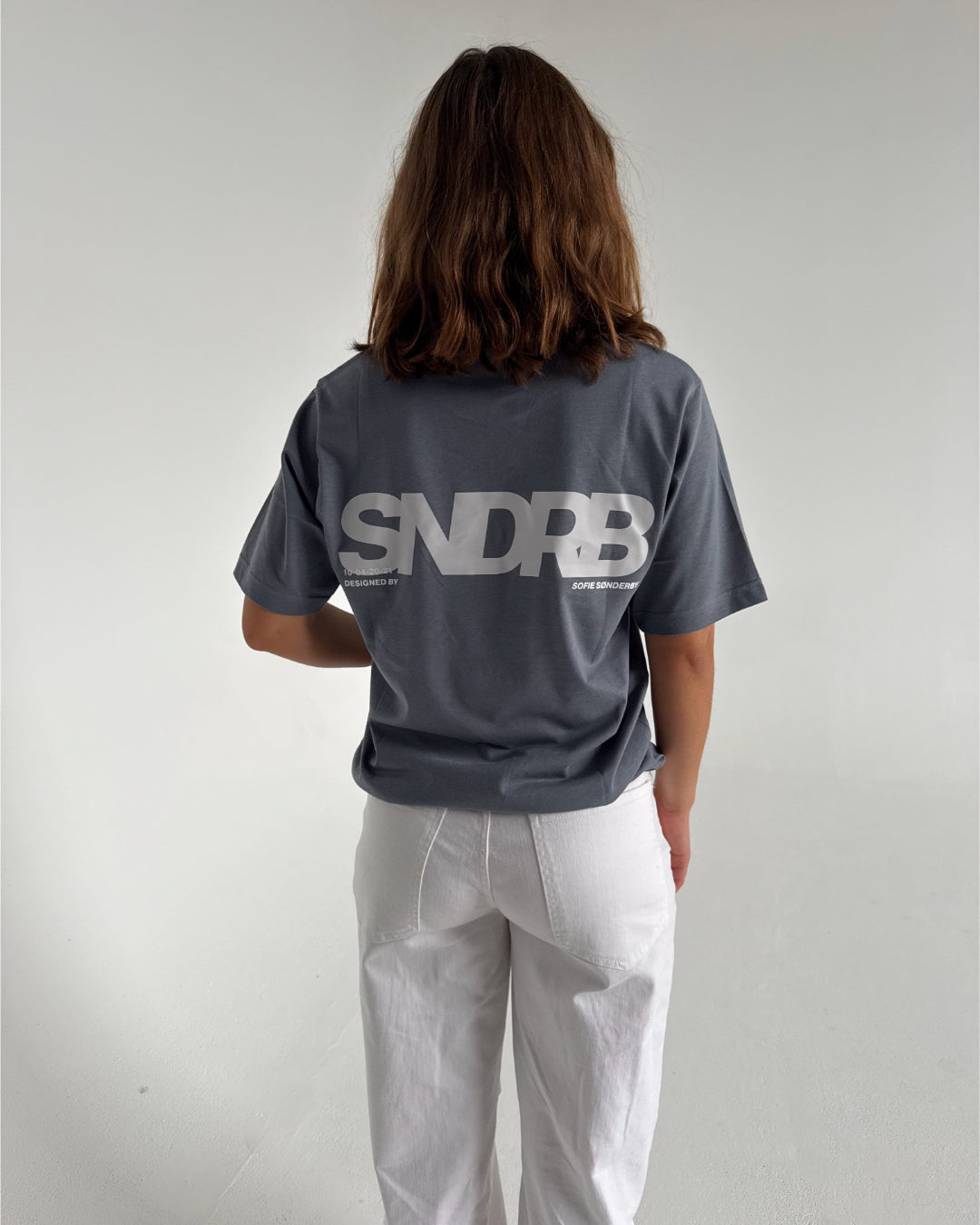 Modelfoto af pige som bærer en SNDRB tee i farven stone, samt et par hvide jeans. Alt fra By Sofie Sønderby
