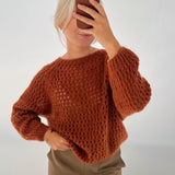 En person iklædt en brun strikket sweater tager en selfie med en mobiltelefon i et lyst rum. De har lyst hår og bærer brune bukser.