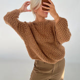 En person tager et spejlfoto iført en brun strikket sweater og beige bukser. Personen holder en telefon op foran ansigtet i et lyst rum.
