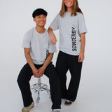 To personer i grå T-shirts med teksten "SØNDERBY". Den ene sidder på en metalstol, den anden står ved siden af. De smiler i et hvidt studie.