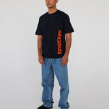 En mand står i afslappet tøj; han bærer en sort t-shirt med ordet "SØNDERBY" skrevet lodret i orange. Han er i et minimalistisk miljø uden distraktioner.