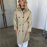 En kvinde står iført en beige trenchcoat med bælte, hvide bukser og sorte sko. Hun er placeret i et fotostudie med beton- og metalbaggrund.
