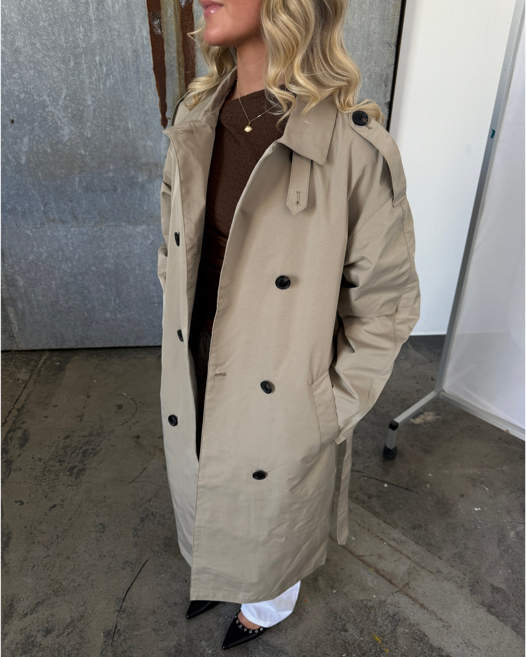 Kvinde iført beige trenchcoat står indendørs på betongulv. Hun har krøllet hår og en brun trøje under frakken. Ved siden af er en metalramme.
