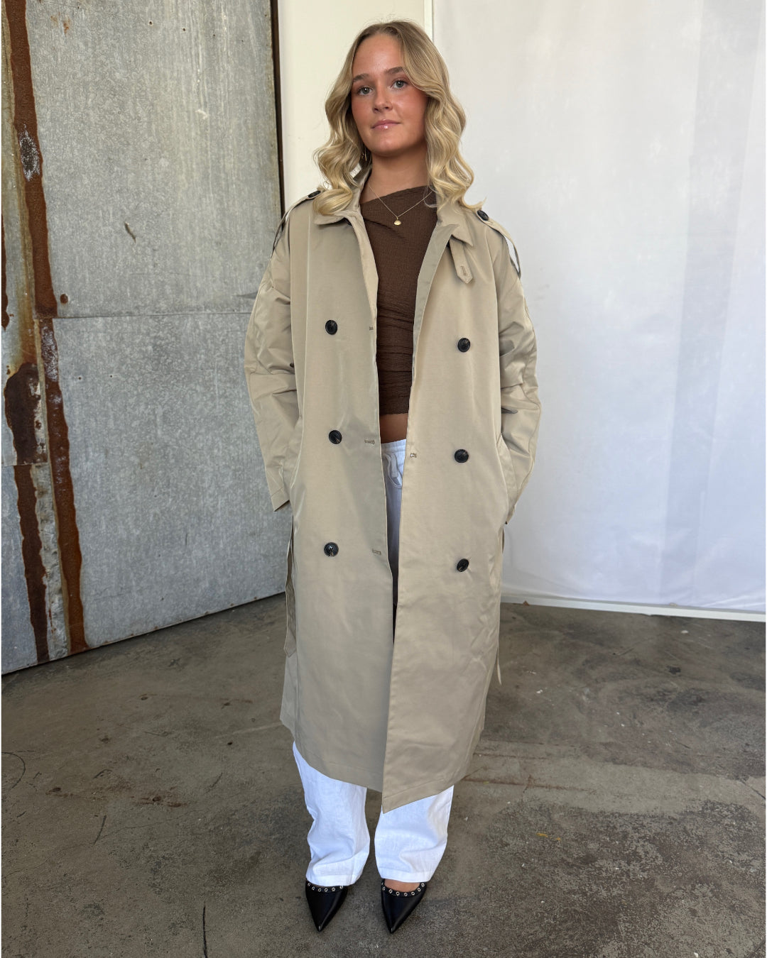 En kvinde står iført en beige trenchcoat, brun trøje og hvide bukser. Hendes hænder er i lommerne. Baggrunden er en rå, industriel væg med beton og metalplader.