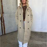 En kvinde står iført en beige trenchcoat, brun trøje og hvide bukser. Hendes hænder er i lommerne. Baggrunden er en rå, industriel væg med beton og metalplader.