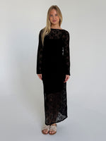 Person i sort blondekjole står stille i et enkelt, lyst rum med hvide vægge. De bærer lyse sandaler og har skulderlangt lyst hår.