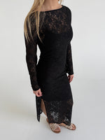 En kvinde iført en sort blondekjole står stille i et hvidt rum, med sandaler på fødderne, og ser nedad.