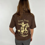 En person iført en brun T-shirt med påtrykt motiv af en cowboy på en hest med teksten "Texas Trails". Personen står mod en neutral, lys baggrund.