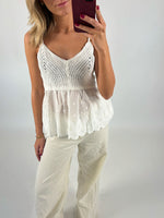 En person iført en hvid, strikket top med blonder, tager en selfie med en rød mobiltelefon i et neutralt lyst rum. Hun har også beige bukser på.