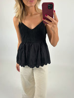 En person iført en sort stropbluse og hvide bukser tager et spejlbillede med en rød telefon i hånden. Baggrunden er neutral og enkel.