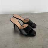 Sort højhælet sandal står stille på en betonoverflade. Sandalen har en enkel rem over foden, åben tå, og stilet hæl. Omgivelsen er minimalistisk med en lys baggrund.