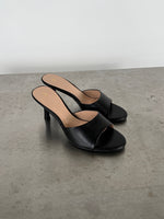 Sort højhælet sandal står stille på en betonoverflade. Sandalen har en enkel rem over foden, åben tå, og stilet hæl. Omgivelsen er minimalistisk med en lys baggrund.