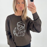 Kvinde står foran et hvidt bagtæppe og tager en selfie med sin telefon. Hun bærer en brun sweatshirt med en hvid minimalistisk tegning af en person, der holder en flaske.