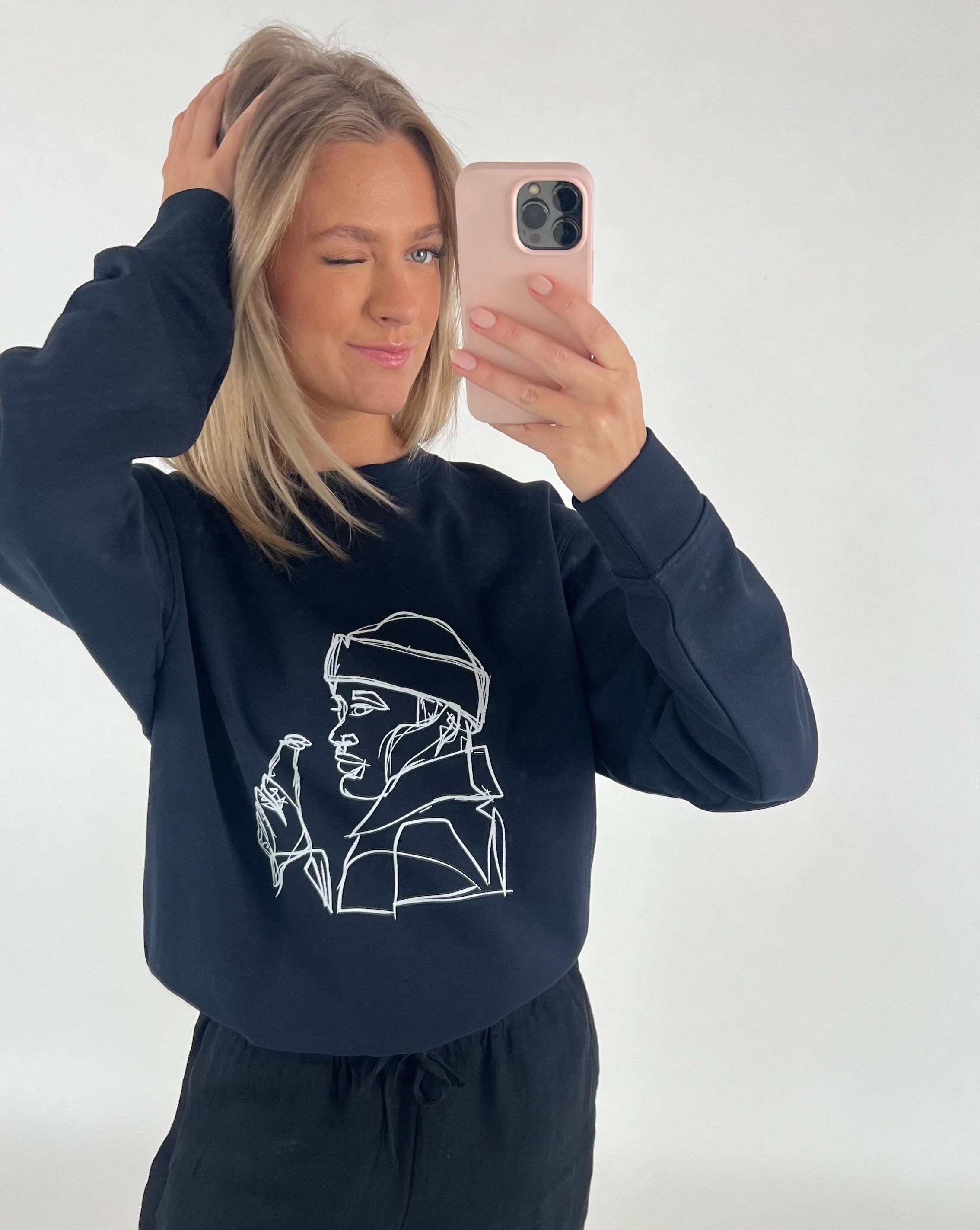 En person tager et selfie med en lyserød telefon og blinker. De bærer en sort trøje med en stregtegning af en person, der holder en flaske.
