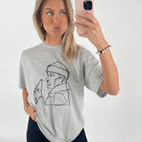 En kvinde fotograferer sig selv med en smartphone, iført en grå T-shirt med en lineart-tegning af en person, der holder en flaske. Hun står i et lyst rum.
