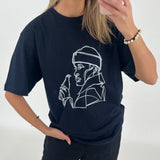 Kvinde iført sort T-shirt med en hvidt lineart design af en person med hætte og flaske. Hun ser sig i spejlet med en mobiltelefon i et lyst rum.